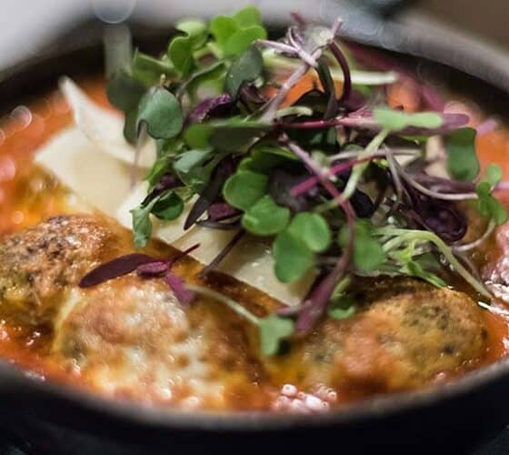 Small bowl with meatballs in a red sauce with shaved parmesan and mixed greens as a topping