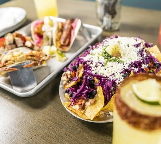 A metal platter holding an assortment of tacos next to a platter of nachos and several glasses of cocktails