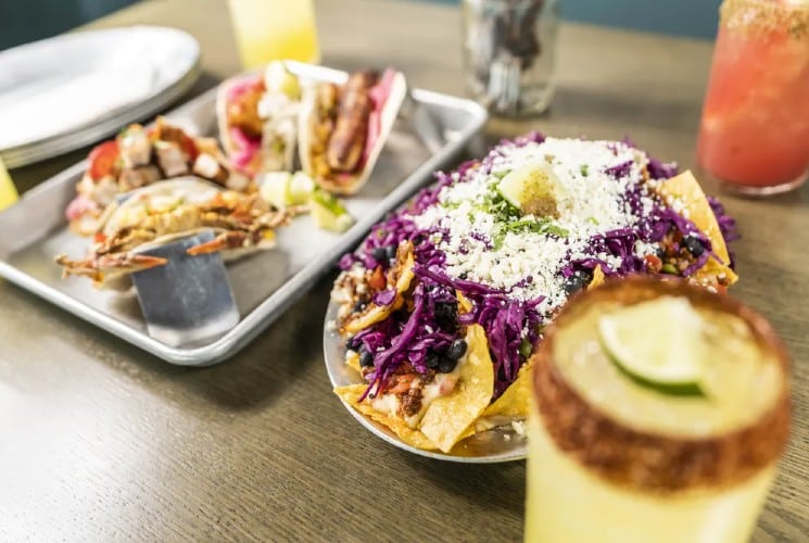 A metal platter holding an assortment of tacos next to a platter of nachos and several glasses of cocktails