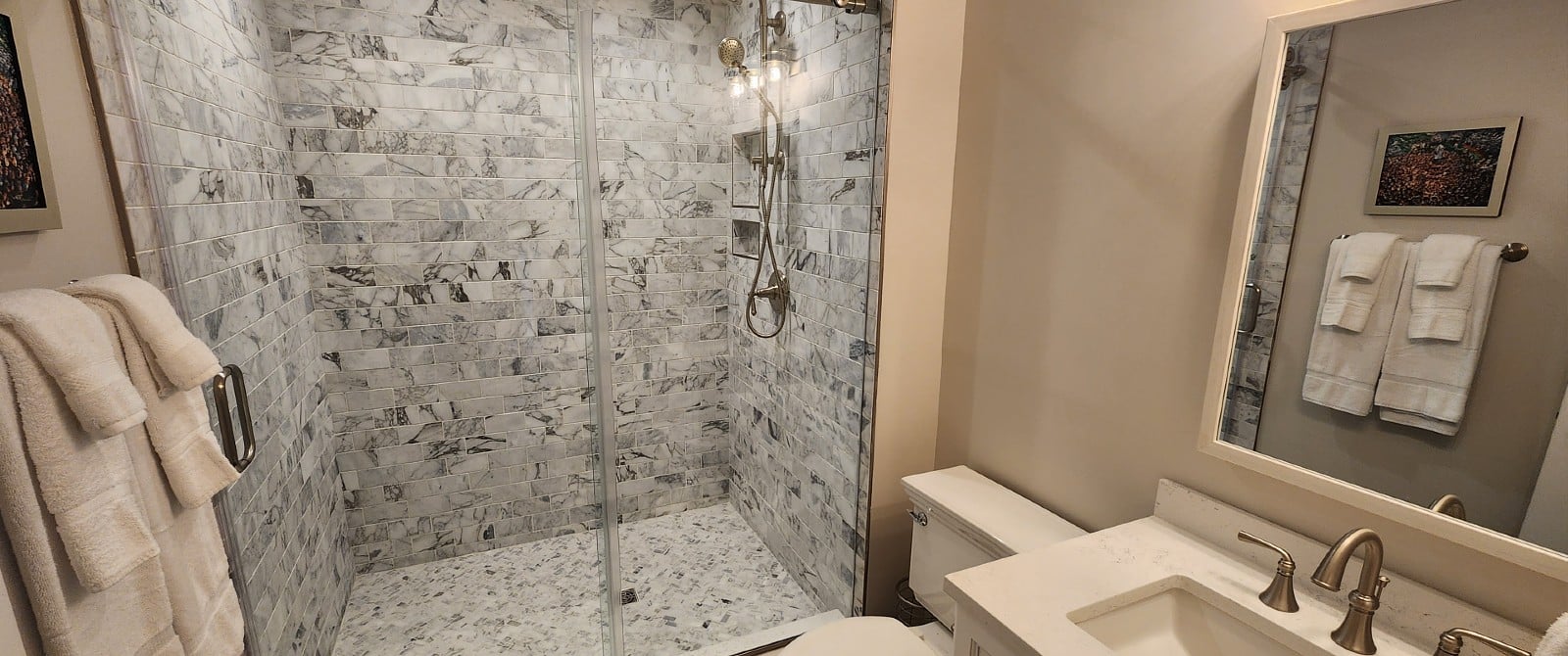 Bathroom with tiled stand up shower with glass doors, toilet, vanity, large mirror and towels hung on the wall