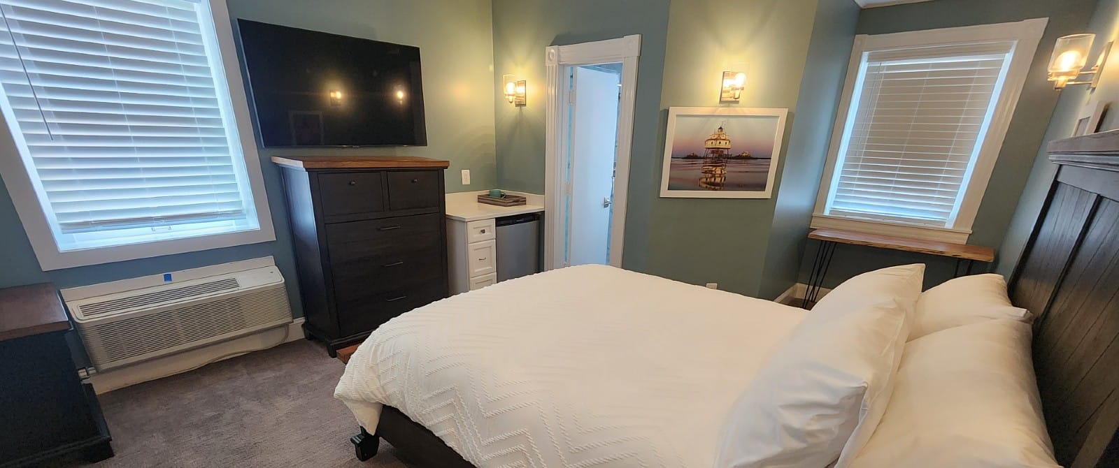 Bedroom with king bed in a white quilt, tall dresser wth TV above, windows with blinds and doorway into a bathroom