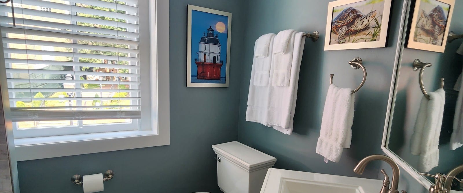 Bathroom with pedestal sink, mirror, white towels, colorful artwork and window with blinds