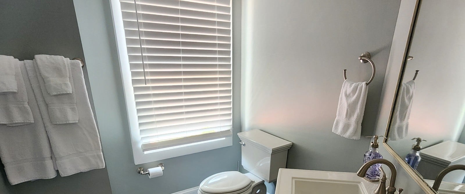 Bathroom with pedestal sink, framed mirror, toilet and window with blinds by hanging towels