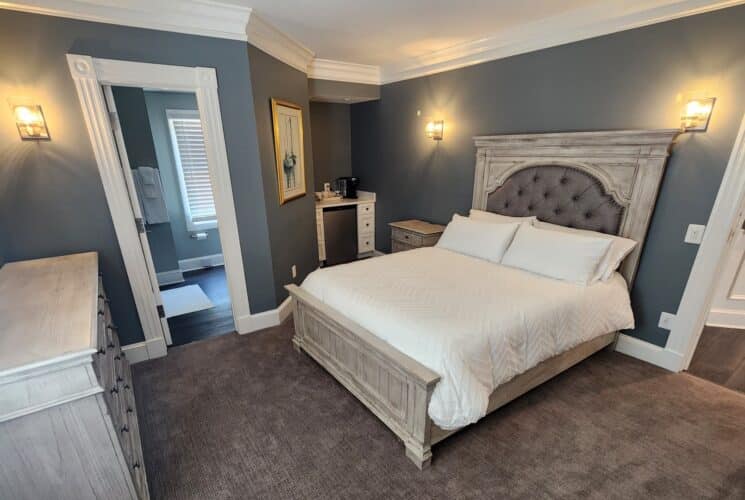 Elegant bedroom in hues of grey with king bed, small kitchenette counter, dresser and doorway into a bathroom
