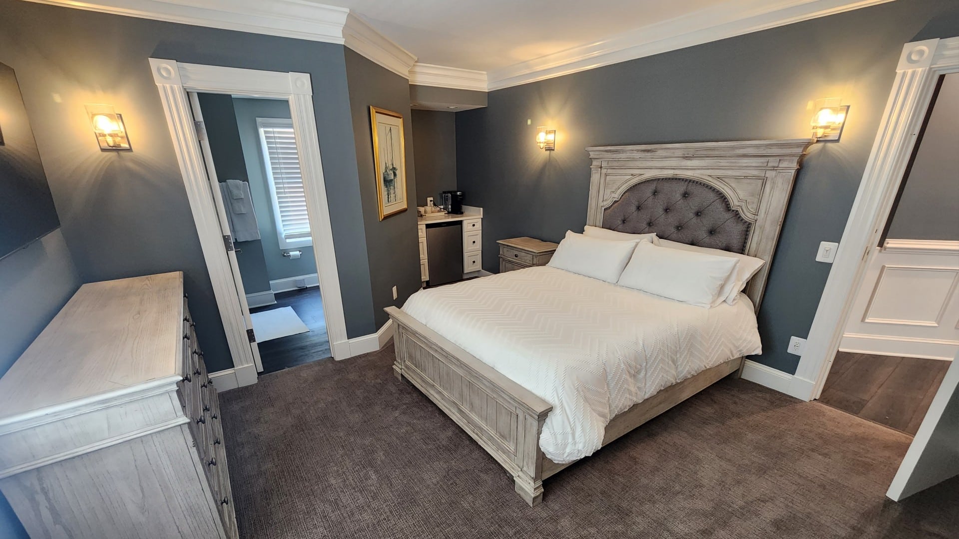 Elegant bedroom in hues of grey with king bed, small kitchenette counter, dresser and doorway into a bathroom