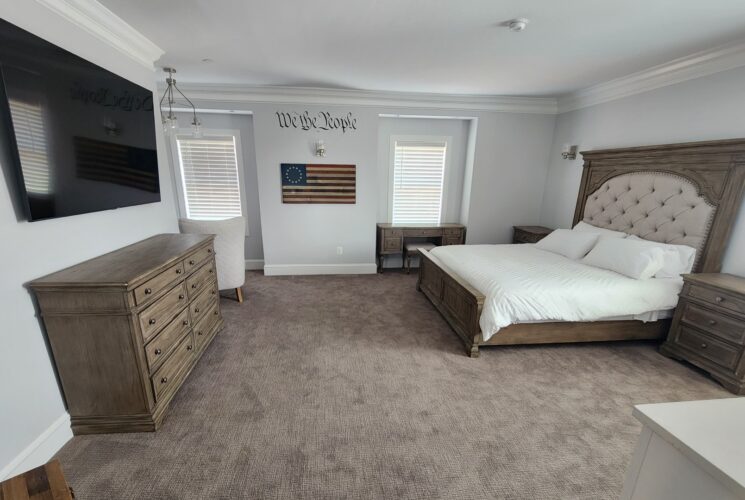 Large bedroom with king bed, TV over a dresser, two side tables and windows with blinds