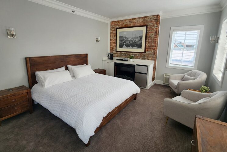 Spacious bedroom with bed, kitchenette counter with brick detail above, two sitting chairs by large windows